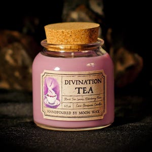 May include: A round glass jar candle with a cork lid. The candle is a light purple color and has a label that reads "Divination Tea." The label features an illustration of a teacup and steam. The candle is hand-poured with moon wax and weighs 6.5 oz.