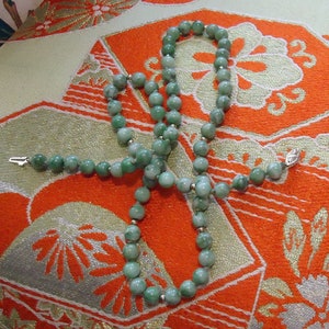May include: A green jade bead necklace with silver clasps. The necklace is laid out on a patterned fabric with orange and white colors.