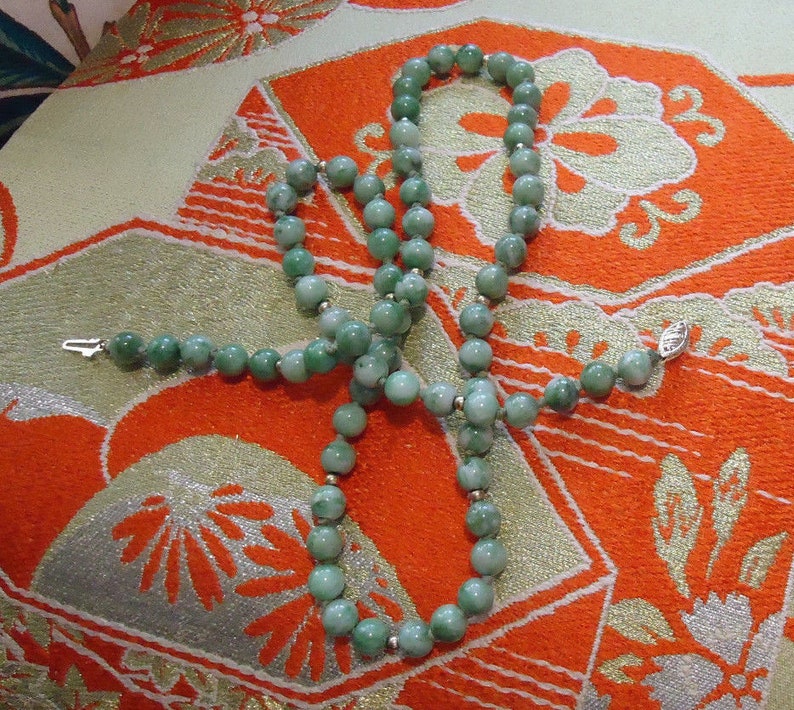 May include: A green jade bead necklace with silver clasps. The necklace is laid out on a patterned fabric with orange and white colors.