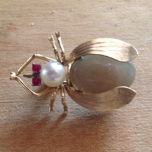 Puede incluir: Un broche de escarabajo dorado con una perla y detalles de rubí. El escarabajo tiene un cuerpo texturizado e iridiscente y alas detalladas.