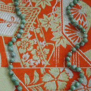 May include: A green jade necklace with gold accents. The necklace is made of round beads and is lying on a patterned fabric.