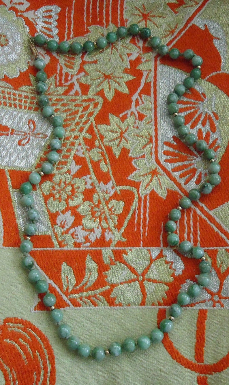 May include: A green jade necklace with gold accents. The necklace is made of round beads and is lying on a patterned fabric.
