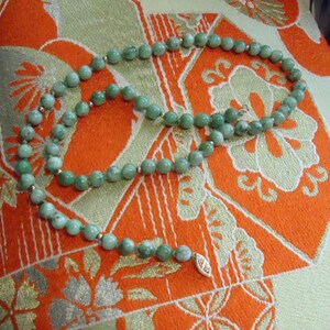 May include: A green jade bead necklace with a gold clasp. The necklace is laid out on a patterned fabric.