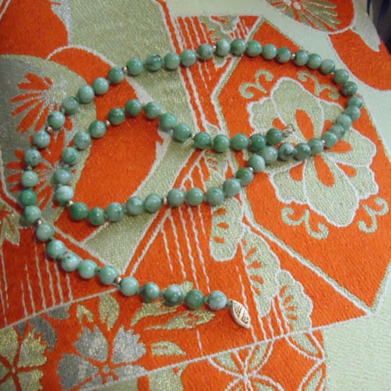 May include: A green jade bead necklace with a gold clasp. The necklace is laid out on a patterned fabric.