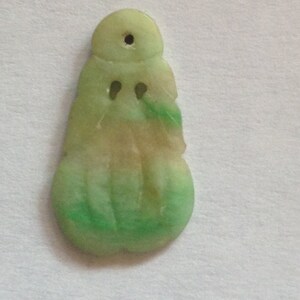 Puede incluir: Un colgante de jade en tonos verde claro y beige. El colgante tiene una parte superior redondeada con un agujero, dos peque&ntilde;os agujeros en forma de ojo y una base bulbosa. El jade tiene un acabado pulido.