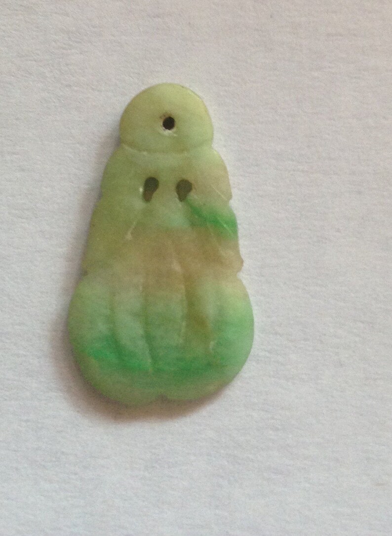 Puede incluir: Un colgante de jade en tonos verde claro y beige. El colgante tiene una parte superior redondeada con un agujero, dos peque&ntilde;os agujeros en forma de ojo y una base bulbosa. El jade tiene un acabado pulido.