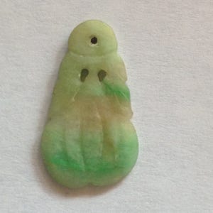 Puede incluir: Un colgante de jade con un dise&ntilde;o tallado. El colgante es principalmente verde con &aacute;reas de color beige y un peque&ntilde;o agujero en la parte superior. La parte inferior tiene una forma redondeada y el dise&ntilde;o general es abstracto.