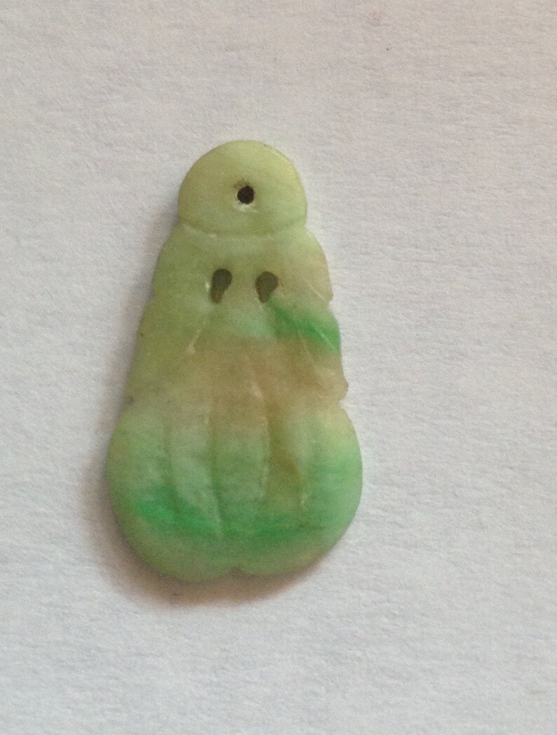 Puede incluir: Un colgante de jade con un dise&ntilde;o tallado. El colgante es principalmente verde con &aacute;reas de color beige y un peque&ntilde;o agujero en la parte superior. La parte inferior tiene una forma redondeada y el dise&ntilde;o general es abstracto.