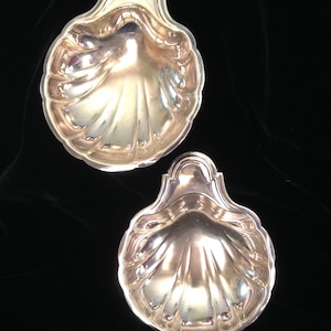 May include: Two silver-colored, shell-shaped dishes. Each dish has a scalloped edge and a decorative handle. The dishes are displayed against a dark background, highlighting their reflective surfaces and intricate design.