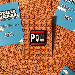 Könnte beinhalten: Eine schwarz-weiße Emaille-Anstecknadel mit dem Wort "POW" in einer Pixel-Schriftart. Die Anstecknadel hat einen roten Rand und ist vor einem Hintergrund aus orangefarbenen und weißen Quadraten platziert.