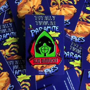 May include: A green, black, and red enamel pin badge with the text "SLIG BARRACKS" on a red banner. The pin is surrounded by a blue background with the text "TOTALLY TUBULAR PARAMITE" in white.