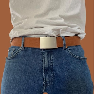 May include: A brown canvas belt with a silver-coloured rectangular buckle is worn with blue jeans and a white t-shirt. The belt is a casual accessory.