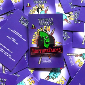 May include: A pin badge with the text "RUPTUREFARMS" in red letters on a black background. The pin features a green alien-like character. The pin is on a purple card with the words "TOTALLY TUBULAR" and "MUDOKON".