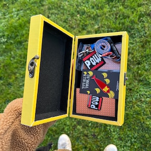 Puede incluir: Una caja amarilla con un interior negro está abierta, revelando una variedad de artículos. La caja contiene un cordón azul con un pin "POW" negro y rojo, un pin de corbata rojo con un diseño de plátano y un pin "POW" negro y naranja. La caja está sentada sobre una superficie verde cubierta de hierba.