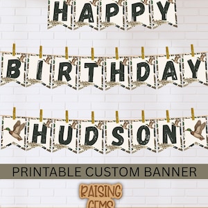 May include: A custom printable banner with the words "HAPPY BIRTHDAY HUDSON" in black letters on individual pennants. Each pennant has a camouflage pattern and a duck illustration. The banner is suspended by clothespins against a white brick wall.