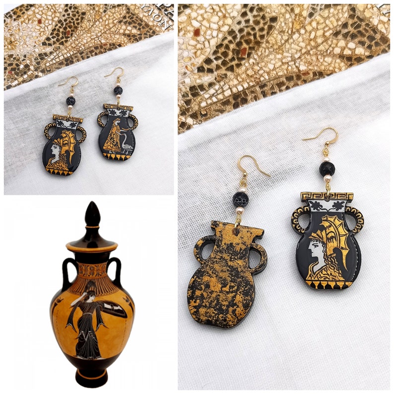 May include: A pair of gold earrings with black and white ceramic amphora pendants. The amphora pendants feature a white silhouette of a woman with a gold crown. The earrings are displayed on a white fabric background.