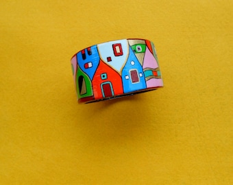 May include: A colorful, hand-painted wooden bracelet featuring a whimsical village scene. The bracelet showcases houses in red, blue, green, and pink with gold accents. The bracelet is approximately 2 inches wide.