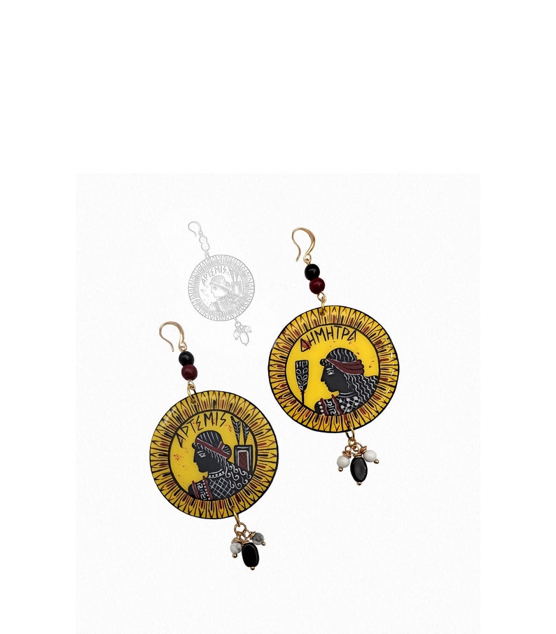 Peut inclure: Une paire de boucles d'oreilles en or avec des pendentifs circulaires jaunes repr&eacute;sentant des illustrations en noir et blanc de visages de femmes. Les pendentifs portent le texte grec "&Alpha;&Rho;&Tau;&Epsilon;&Mu;&Iota;&Sigma;" et "&Delta;&Eta;&Mu;&Eta;&Tau;&Rho;&Alpha;". Les boucles d'oreilles ont des perles noires et blanches qui pendent.