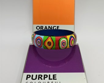 May include: A colourful bangle bracelet with a blue base and a pattern of circles, diamonds, and stripes in red, yellow, green, pink, and white. The bracelet is sitting on a purple surface with the words "PURPLE" and "COLOURFUL" printed in white.