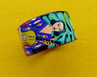 Puede incluir: Un brazalete de madera con un diseño pintado de colores de una mujer tocando una guitarra. La mujer lleva un vestido morado y tiene el pelo negro. El fondo es un patrón de remolino verde azulado y verde.
