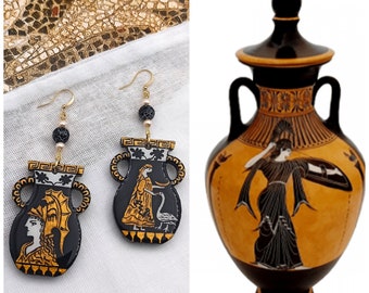 May include: A pair of black and gold earrings shaped like ancient Greek urns with painted figures. The earrings are hanging from gold chains with black beads. The urns have a white and gold design with a figure of a woman and a bird.