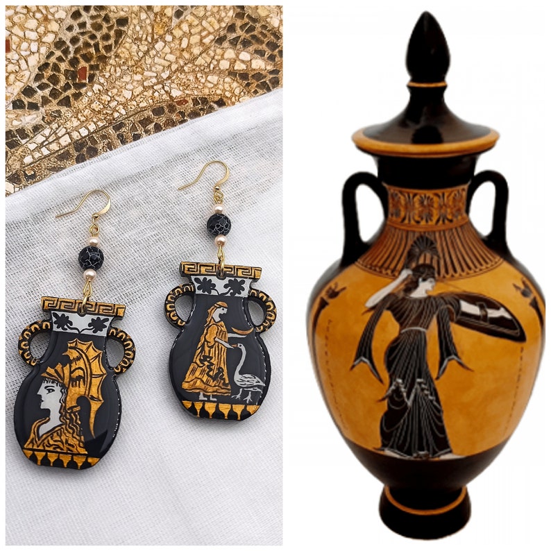 May include: A pair of black and gold earrings shaped like ancient Greek urns with painted figures. The earrings are hanging from gold chains with black beads. The urns have a white and gold design with a figure of a woman and a bird.