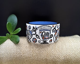Puede incluir: Un brazalete de cerámica blanca con un diseño de llave griega azul, negro y naranja. El brazalete presenta una imagen estilizada de un jarrón con un pájaro en la parte superior.