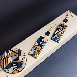 Può includere: Un set di orecchini geometrici e una spilla con un motivo blu, rosa e oro. Gli orecchini sono orecchini pendenti con una perla blu e un gancio dorato.