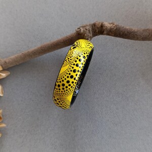 May include: A yellow and black bangle bracelet with a dotted pattern. The bracelet is resting on a brown branch against a gray background. The bracelet has a glossy finish and a black interior.