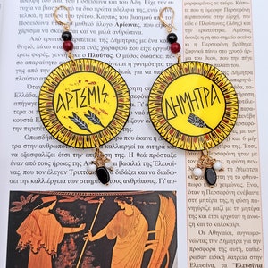 Peut inclure: Une paire de boucles d'oreilles circulaires jaunes et rouges avec des anneaux dor&eacute;s. Les boucles d'oreilles pr&eacute;sentent des perles noires et blanches et les mots grecs "Artemis" et "D&eacute;m&eacute;ter" &eacute;crits en noir.