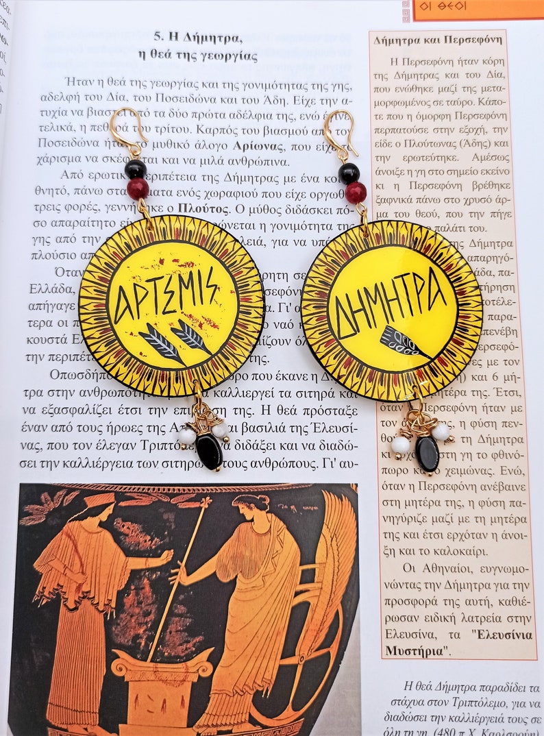 Peut inclure: Une paire de boucles d'oreilles circulaires jaunes et rouges avec des anneaux dor&eacute;s. Les boucles d'oreilles pr&eacute;sentent des perles noires et blanches et les mots grecs "Artemis" et "D&eacute;m&eacute;ter" &eacute;crits en noir.
