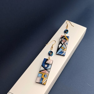 May include: A pair of geometric patterned earrings with blue, yellow, pink, and white colours. The earrings are made of metal and have a gold finish. Each earring features a blue bead and a rectangular pendant with a geometric pattern.