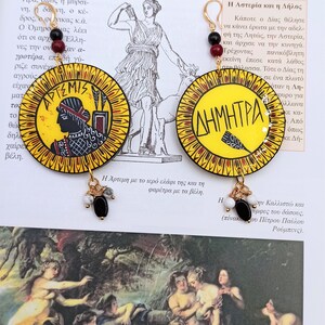 Peut inclure: Une paire de boucles d'oreilles cr&eacute;oles dor&eacute;es avec un pendentif circulaire jaune, rouge et noir repr&eacute;sentant la d&eacute;esse grecque Art&eacute;mis et le texte "&Alpha;&Rho;&Tau;&Epsilon;&Mu;&Iota;&Sigma;". L'autre boucle d'oreille repr&eacute;sente la d&eacute;esse grecque D&eacute;m&eacute;ter et le texte "&Delta;&Eta;&Mu;&Eta;&Tau;&Rho;&Alpha;".
