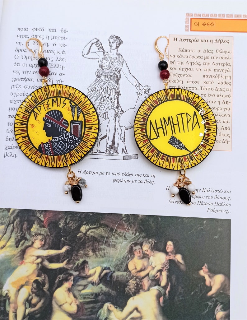 Peut inclure: Une paire de boucles d'oreilles cr&eacute;oles dor&eacute;es avec un pendentif circulaire jaune, rouge et noir repr&eacute;sentant la d&eacute;esse grecque Art&eacute;mis et le texte "&Alpha;&Rho;&Tau;&Epsilon;&Mu;&Iota;&Sigma;". L'autre boucle d'oreille repr&eacute;sente la d&eacute;esse grecque D&eacute;m&eacute;ter et le texte "&Delta;&Eta;&Mu;&Eta;&Tau;&Rho;&Alpha;".