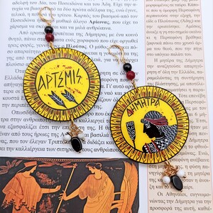 Peut inclure: Une paire de boucles d'oreilles cr&eacute;oles dor&eacute;es avec des pendentifs ronds peints en jaune, rouge et noir. Les pendentifs repr&eacute;sentent les d&eacute;esses grecques Art&eacute;mis et D&eacute;m&eacute;ter avec leurs noms &eacute;crits en grec.