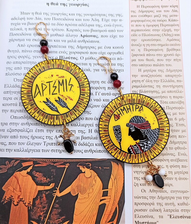 Peut inclure: Une paire de boucles d'oreilles cr&eacute;oles dor&eacute;es avec des pendentifs ronds peints en jaune, rouge et noir. Les pendentifs repr&eacute;sentent les d&eacute;esses grecques Art&eacute;mis et D&eacute;m&eacute;ter avec leurs noms &eacute;crits en grec.