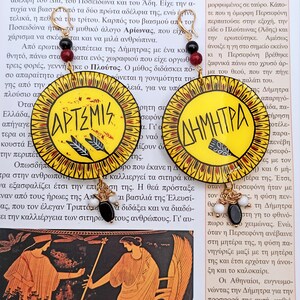 Peut inclure: Une paire de boucles d'oreilles rondes en bois peintes en jaune, rouge et noir avec des accents dor&eacute;s. Les boucles d'oreilles pr&eacute;sentent les mots "Artemis" et "D&eacute;m&eacute;ter" en lettres noires.
