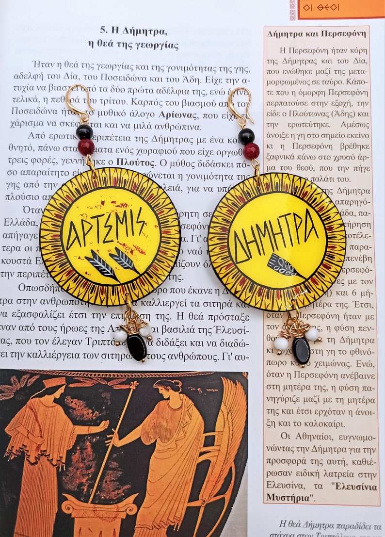 Peut inclure: Une paire de boucles d'oreilles rondes en bois peintes en jaune, rouge et noir avec des accents dor&eacute;s. Les boucles d'oreilles pr&eacute;sentent les mots "Artemis" et "D&eacute;m&eacute;ter" en lettres noires.