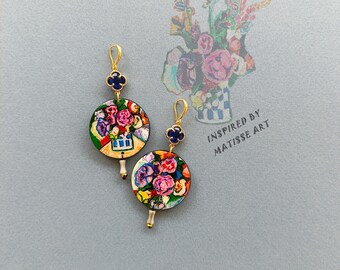 May include: A pair of gold earrings with a blue clover charm and a round, colourful floral design inspired by Matisse art.  INSPIRED BY MATISSE ART is written on the background.