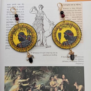 Peut inclure: Une paire de boucles d'oreilles rondes, jaunes et rouges avec des perles noires et blanches qui pendent. Les boucles d'oreilles pr&eacute;sentent des images peintes de d&eacute;esses grecques, Art&eacute;mis et D&eacute;m&eacute;ter, avec leurs noms &eacute;crits en lettres grecques.
