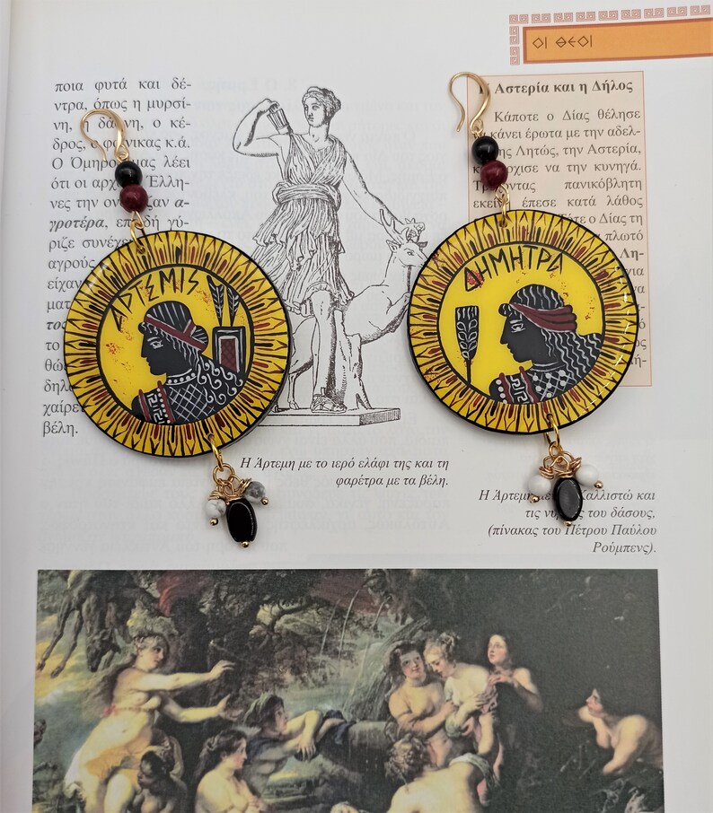 Peut inclure: Une paire de boucles d'oreilles rondes, jaunes et rouges avec des perles noires et blanches qui pendent. Les boucles d'oreilles pr&eacute;sentent des images peintes de d&eacute;esses grecques, Art&eacute;mis et D&eacute;m&eacute;ter, avec leurs noms &eacute;crits en lettres grecques.