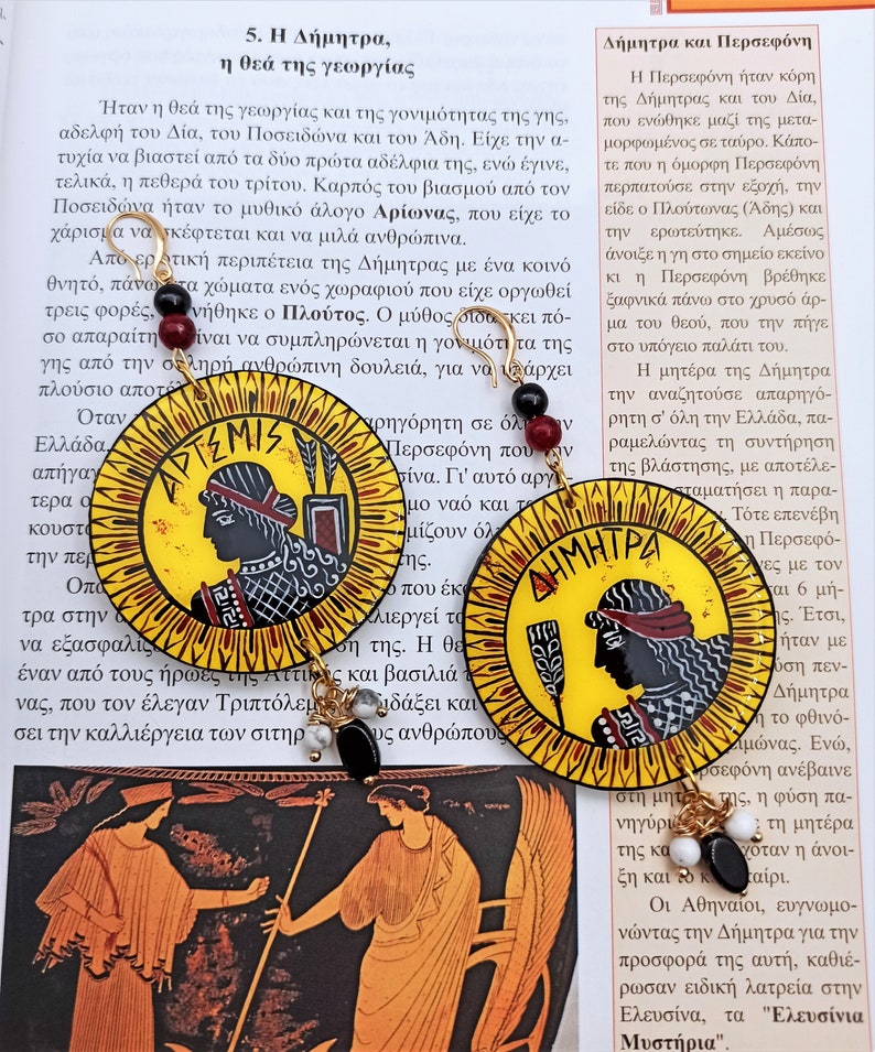 Peut inclure: Deux boucles d'oreilles rondes peintes en jaune et noir avec des accents dor&eacute;s. Les boucles d'oreilles pr&eacute;sentent des images des d&eacute;esses grecques Art&eacute;mis et D&eacute;m&eacute;ter. Les textes "&Alpha;&Rho;&Tau;&Epsilon;&Mu;&Iota;&Sigma;" et "&Delta;&Eta;&Mu;&Eta;&Tau;&Rho;&Alpha;" sont visibles sur les boucles d'oreilles.