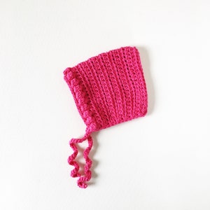 May include: A pink crocheted baby bonnet with a long, curled tie. The bonnet has a textured design and is made with a soft yarn.