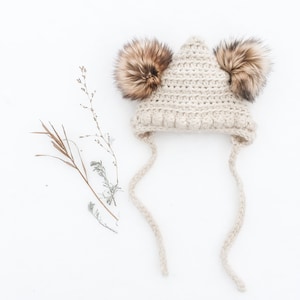 May include: A cream-colored crocheted hat with two fluffy brown pom-poms on top. The hat has long ties that hang down on either side.