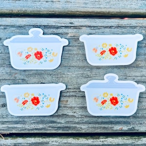 May include: Four stickers of light blue casserole dishes with lids, adorned with floral patterns in red, yellow, and blue. The stickers are arranged on a weathered wooden surface, showcasing a vintage kitchenware theme.