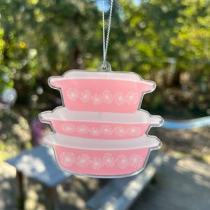 May include: A pink and white glass ornament shaped like a stack of three vintage casserole dishes. The dishes have a white daisy pattern around the rim.