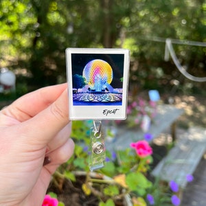 May include: A clear plastic badge holder with a photo of the Spaceship Earth attraction at Epcot. The photo is in a white frame with the word "Epcot" printed below it. The badge holder has a silver retractable clip.