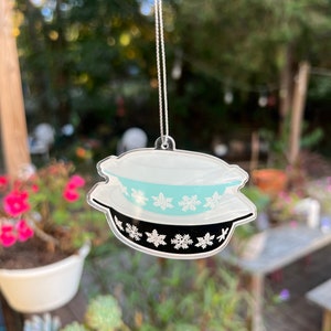 May include: A turquoise and white ceramic bowl ornament with a black rim and snowflake design. The ornament is hanging from a silver string.