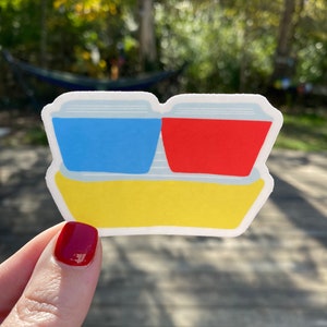 May include: A sticker of two stacked containers, one blue and one red, with a yellow base. The sticker is on a wooden surface.