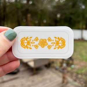 May include: A white sticker with a yellow floral design. The design features a central flower with four petals and leaves on either side.